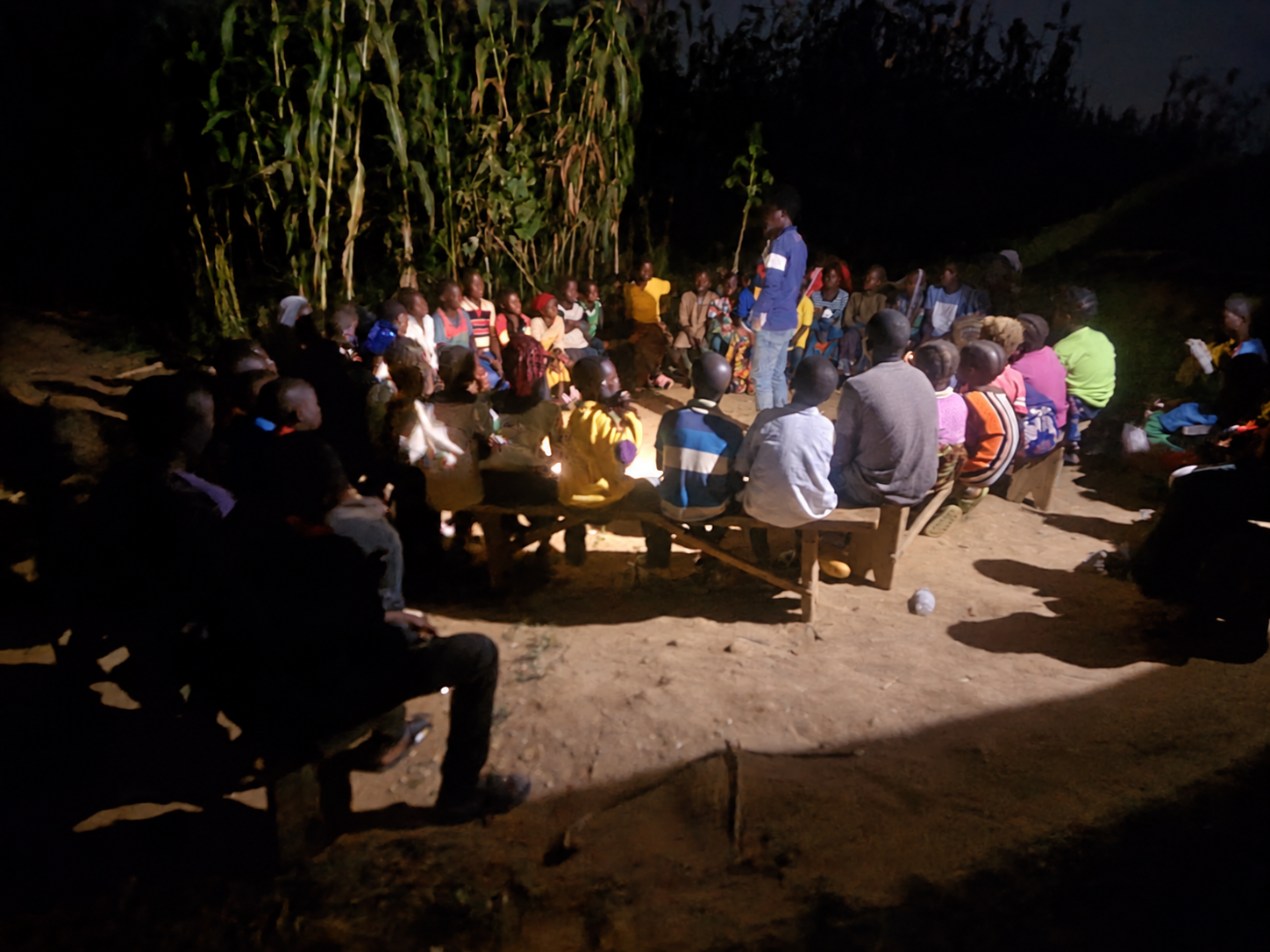 People gathered on benches outdoors at night, with a person standing among them.